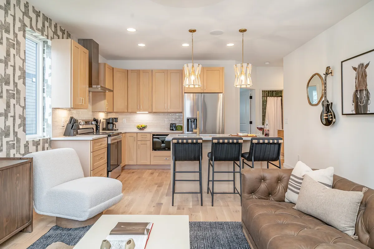 A contemporary space featuring a kitchen island with elegant bar stools, a tufted chocolate brown sofa, decorative wall art, and warm hanging light fixtures. The kitchen blends into the living area, promoting a social and connected environment ideal for group meals and conversation.