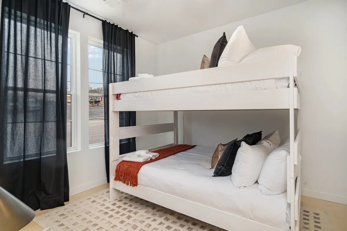 A cozy bunk room with a sturdy bunk bed, large windows draped with black curtains, and decorative pillows. The space feels playful yet restful, suitable for children or friends sharing a room.