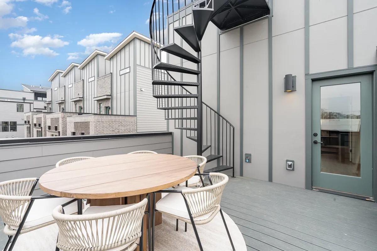 A modern rooftop patio featuring a spiral staircase, a spacious dining table, and comfortable outdoor seating. The sky is visible above, suggesting an open and inviting space for group gatherings and alfresco dining.