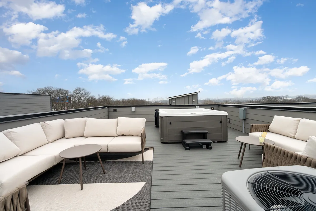 The image showcases a stylish rooftop lounge area with plush sofa seating, a bubbling hot tub, and a chic outdoor rug, all set beneath an open sky with the Nashville skyline visible in the background. The scene is inviting, with ample space for groups to relax, socialize, and enjoy sunset views, suggesting an ideal setting for gathering after a day of exploring the city.