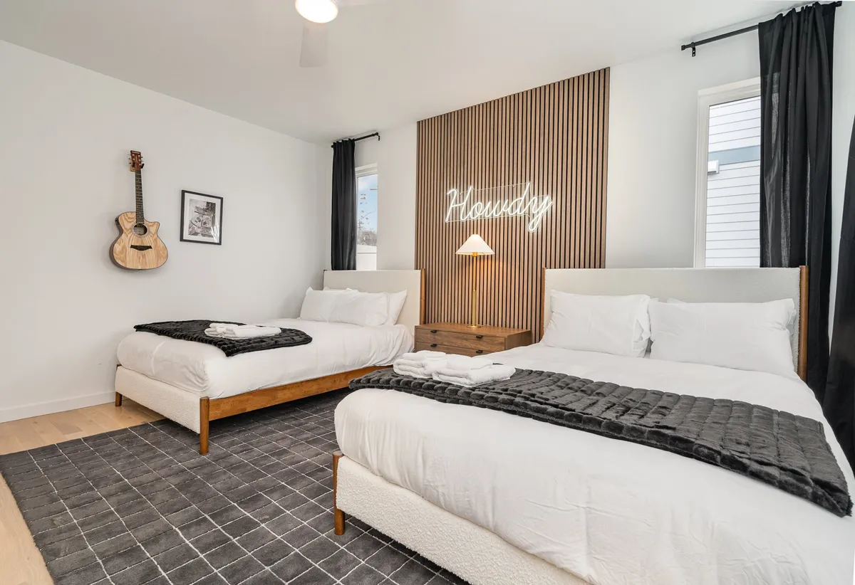 A modern bedroom with two plush beds, vibrant bedding, and a glowing neon sign reading 'Housdzy.' An acoustic guitar hangs on the wall, adding a playful touch. The room feels inviting and is perfect for group stays or families.