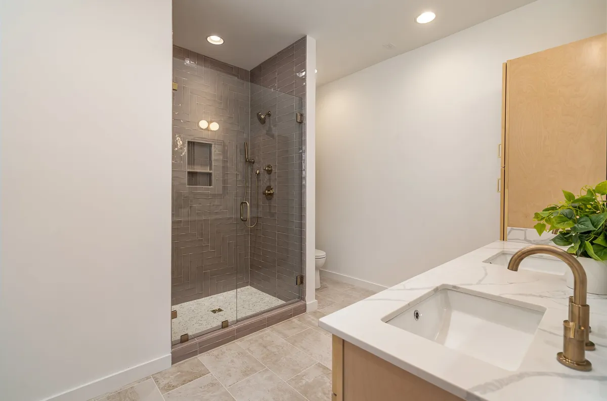 A modern bathroom featuring a glass shower enclosure, marble countertop, and warm wooden cabinetry. The space looks clean, spacious, and spa-inspired, ideal for rejuvenation.